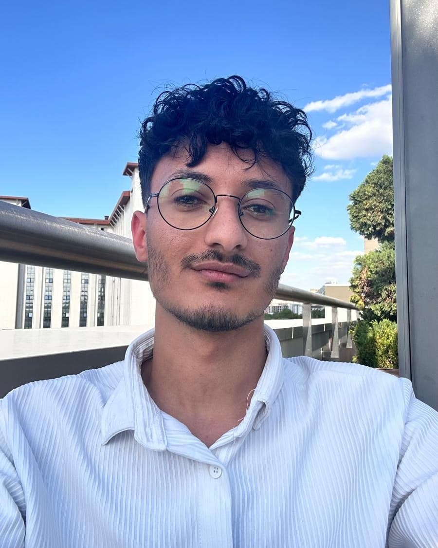 Yaaqob Alfakih, wearing round glasses and a white shirt, photographed outdoors on a balcony.